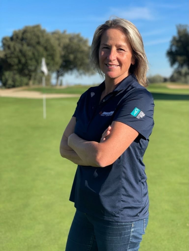 Mujer con los brazos cruzados, en un campo de golf, vistiendo un polo azul marino y jeans, sonriendo.