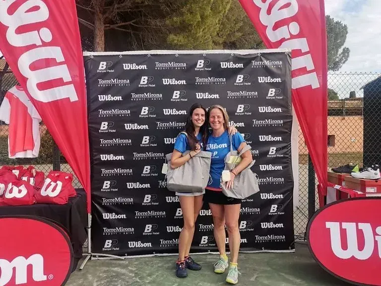 Dos mujeres sonríen, sosteniendo bolsas frente a pancartas de Wilson en un evento de tenis al aire libre.