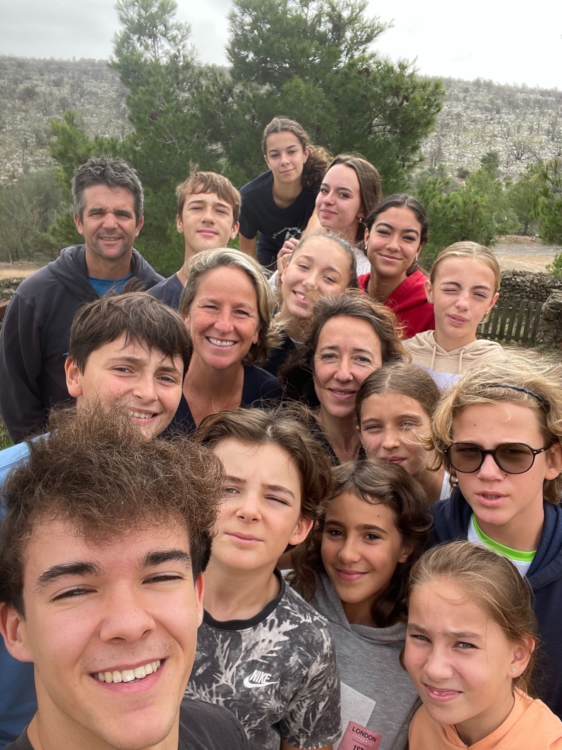 Selfie grupal al aire libre; grupo diverso sonriendo a la cámara. Día nublado, piel clara, fondo natural.
