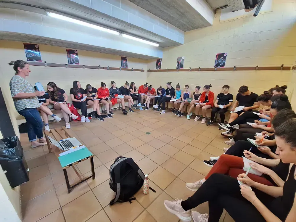 Una mujer da una conferencia a un grupo de jóvenes sentados en círculo en una sala con carteles.