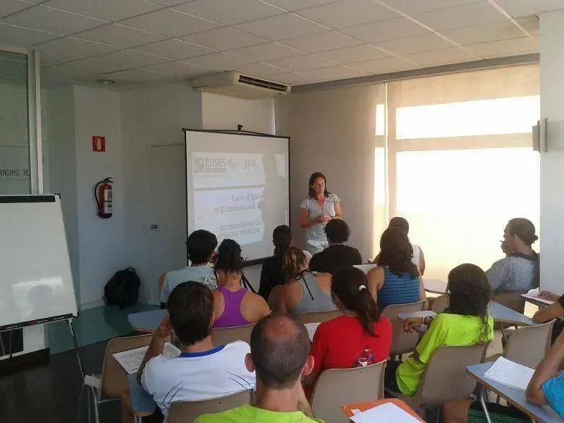 Una mujer da una presentación a una audiencia sentada en un aula bien iluminada.