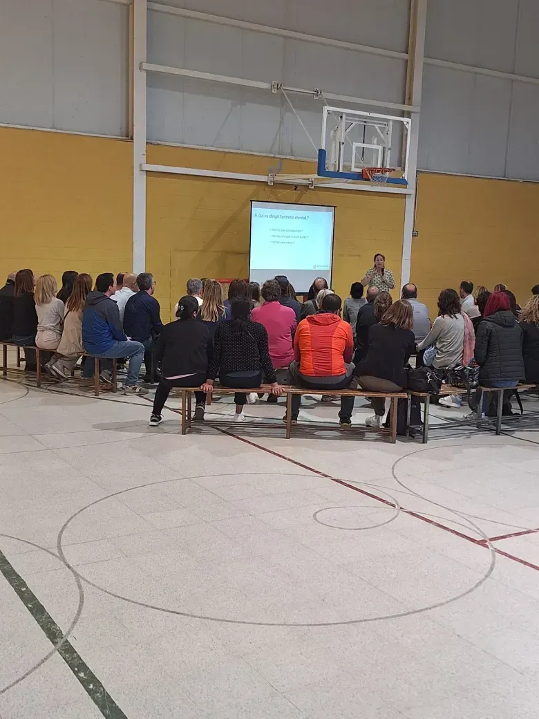 Público sentado, escuchando una presentación en un gimnasio; un hombre de pie junto a la pantalla.
