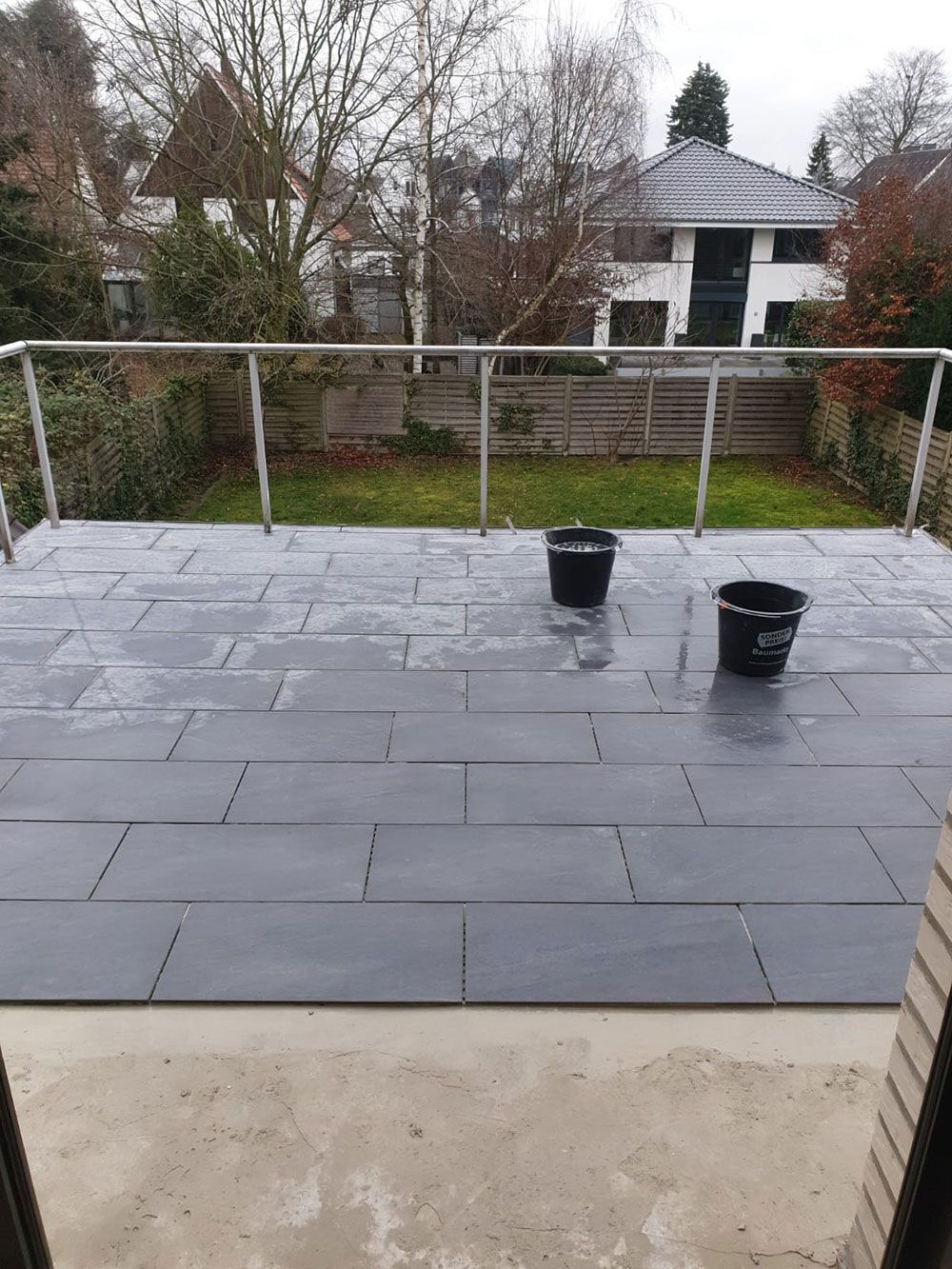 Terrasse mit grauen rechteckigen Fliesen, zwei schwarzen Eimern und einem Metallgeländer mit Blick auf einen Hof.