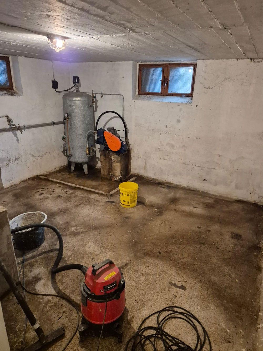 Keller mit einem Warmwasserbereiter, einem Staubsauger und einem gelben Eimer auf einem schmutzigen Betonboden.