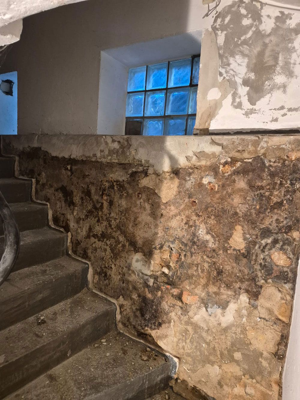 Treppenhaus mit beschädigter, fleckiger Wand. Darüber befindet sich ein Fenster mit blau getönten Glasbausteinen.