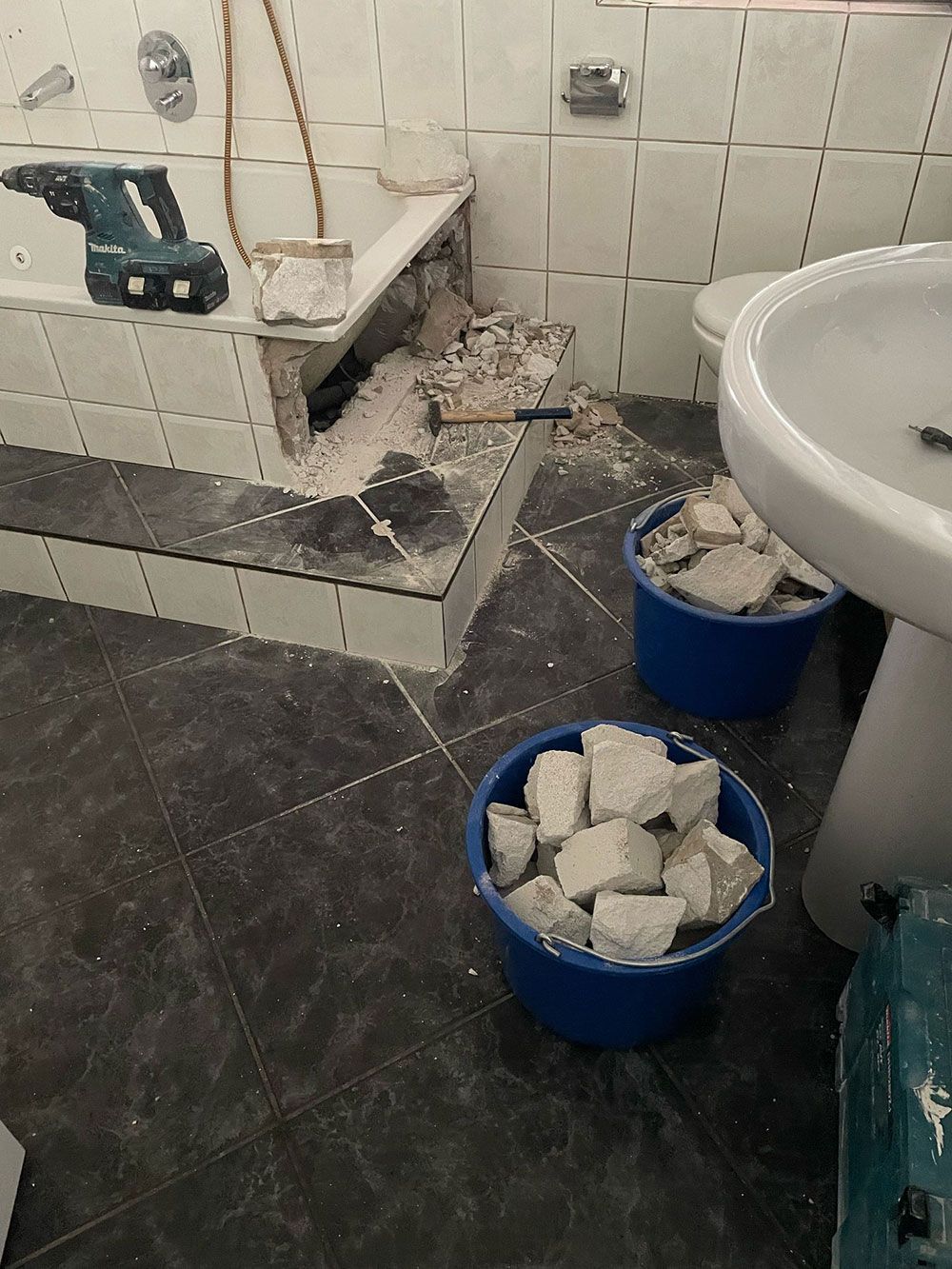 Badezimmerrenovierung im Gange: mit Schutt gefüllte Eimer, freiliegendes Wanneninneres und Bohrer auf gefliestem Sims.