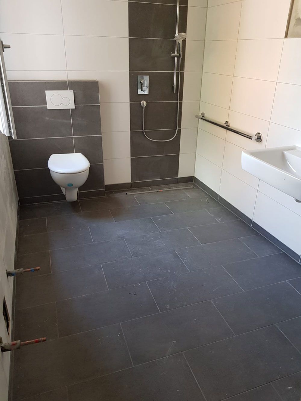 Barrierefreies Badezimmer mit dunkelgrauen Bodenfliesen, weißen und grauen Wandfliesen, Toilette, Dusche und Haltegriffen.