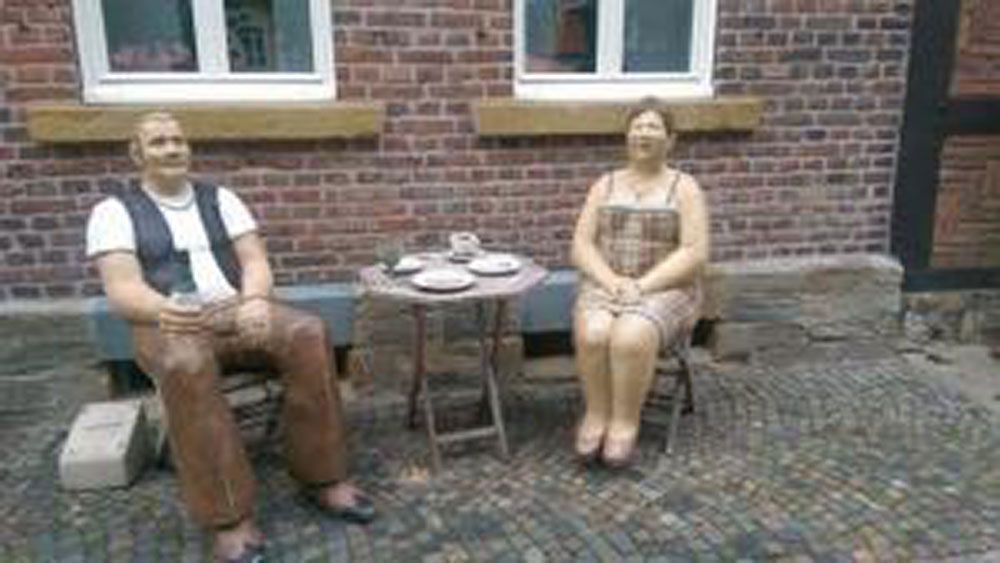 Statuen eines Mannes und einer Frau, die vor einem Backsteingebäude an einem Tisch mit Tee sitzen.