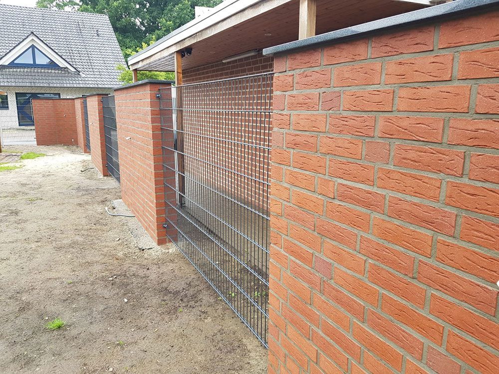 Backsteinmauer mit Maschendrahtzaunabschnitt; Wohnumgebung mit rotem Backstein und hellbraunem Dach.