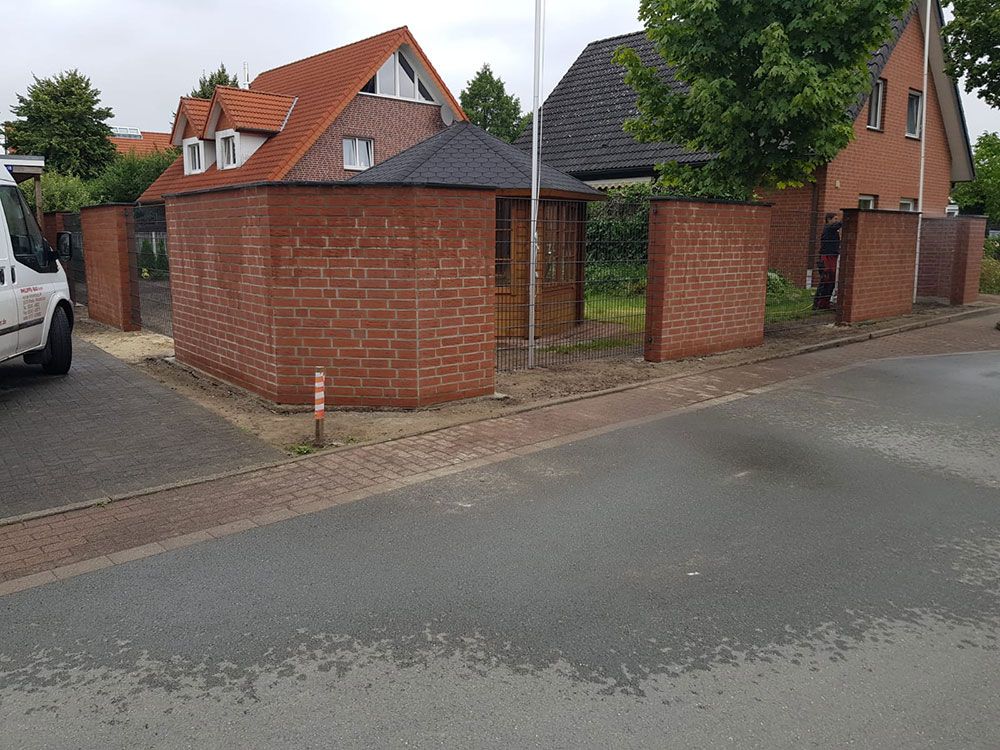 Rote Backsteinmauer umgibt einen kleinen Garteneingang an einer Straße mit einem geparkten Lieferwagen und Häusern.