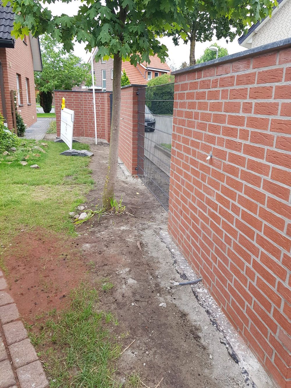 Eine Backsteinmauer grenzt an eine Rasenfläche und einen Feldweg. In der Nähe wächst ein Baum.