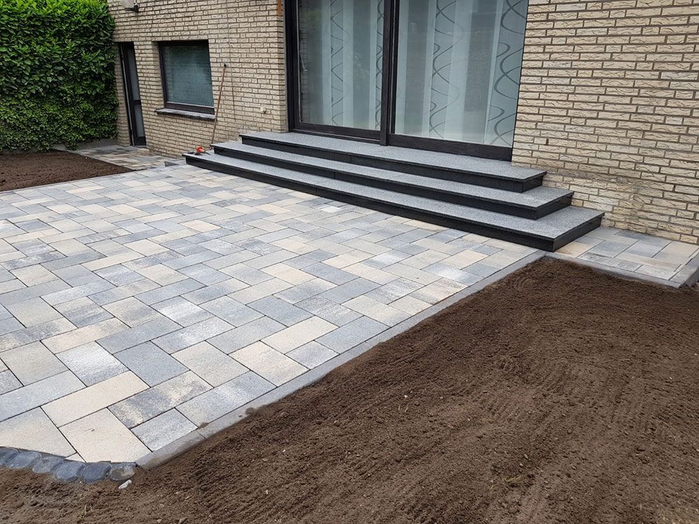 Eine Terrasse mit grauen und beigen Pflastersteinen, Stufen führen zu einer Glastür, neben einem Backsteingebäude.