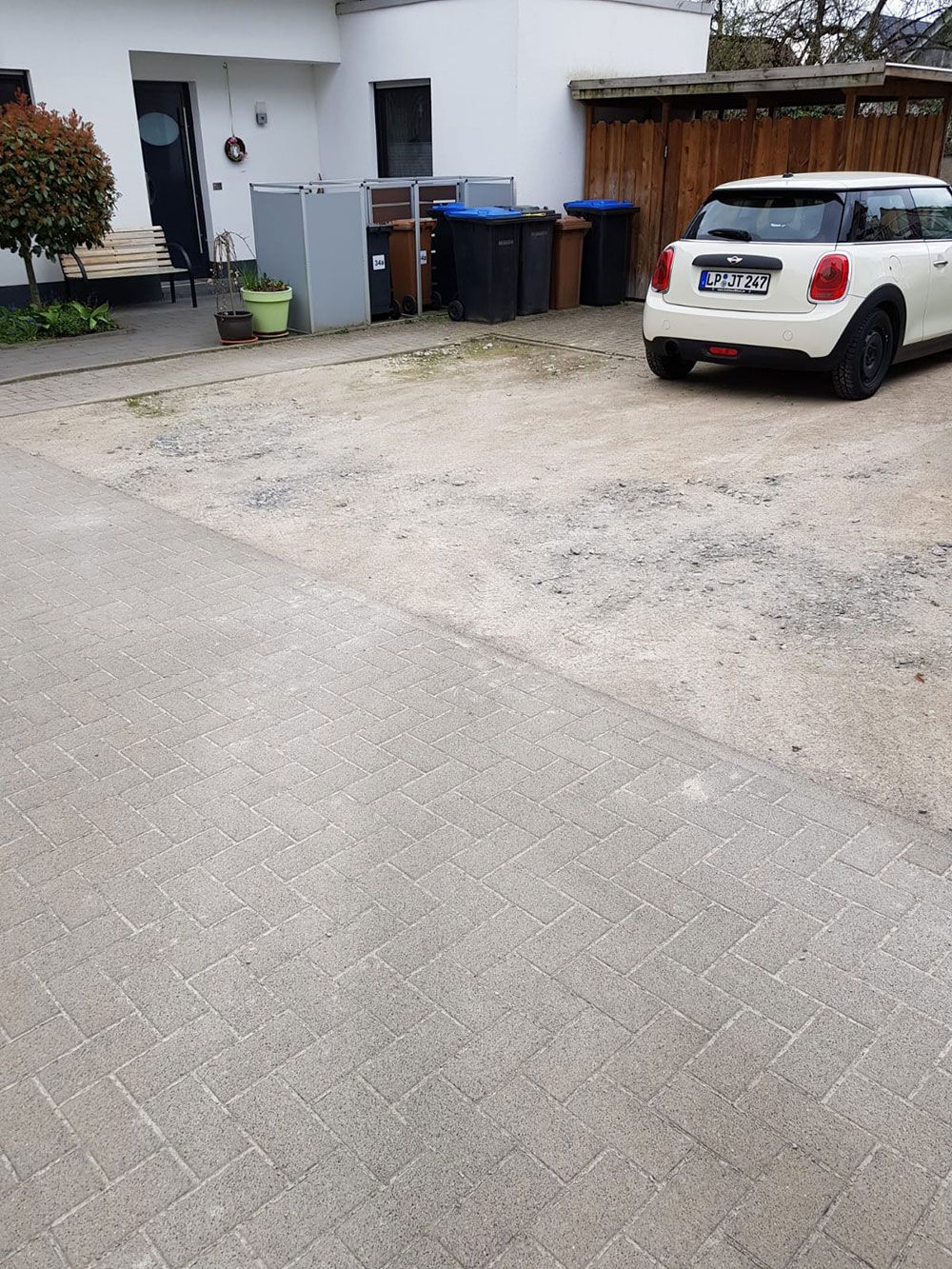 Schotterparkplatz mit einem weißen Auto, Mülltonnen und einem Haus im Hintergrund.