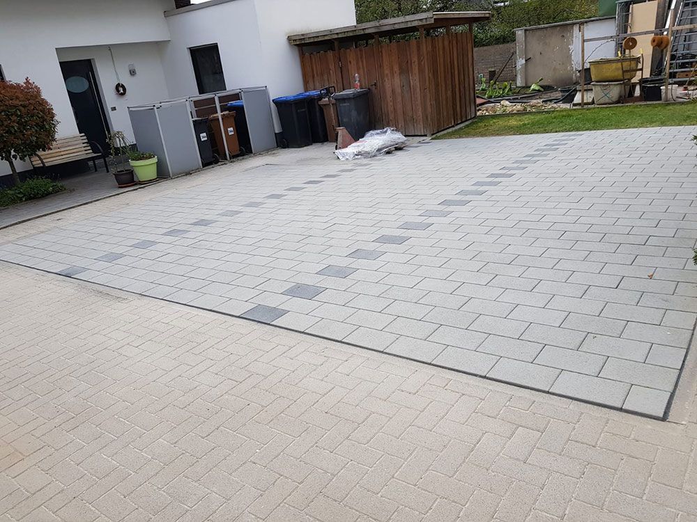 Gepflasterte Terrasse mit dekorativer Umrandung. Im Hintergrund sind ein Schuppen, Mülltonnen und ein Haus zu sehen.