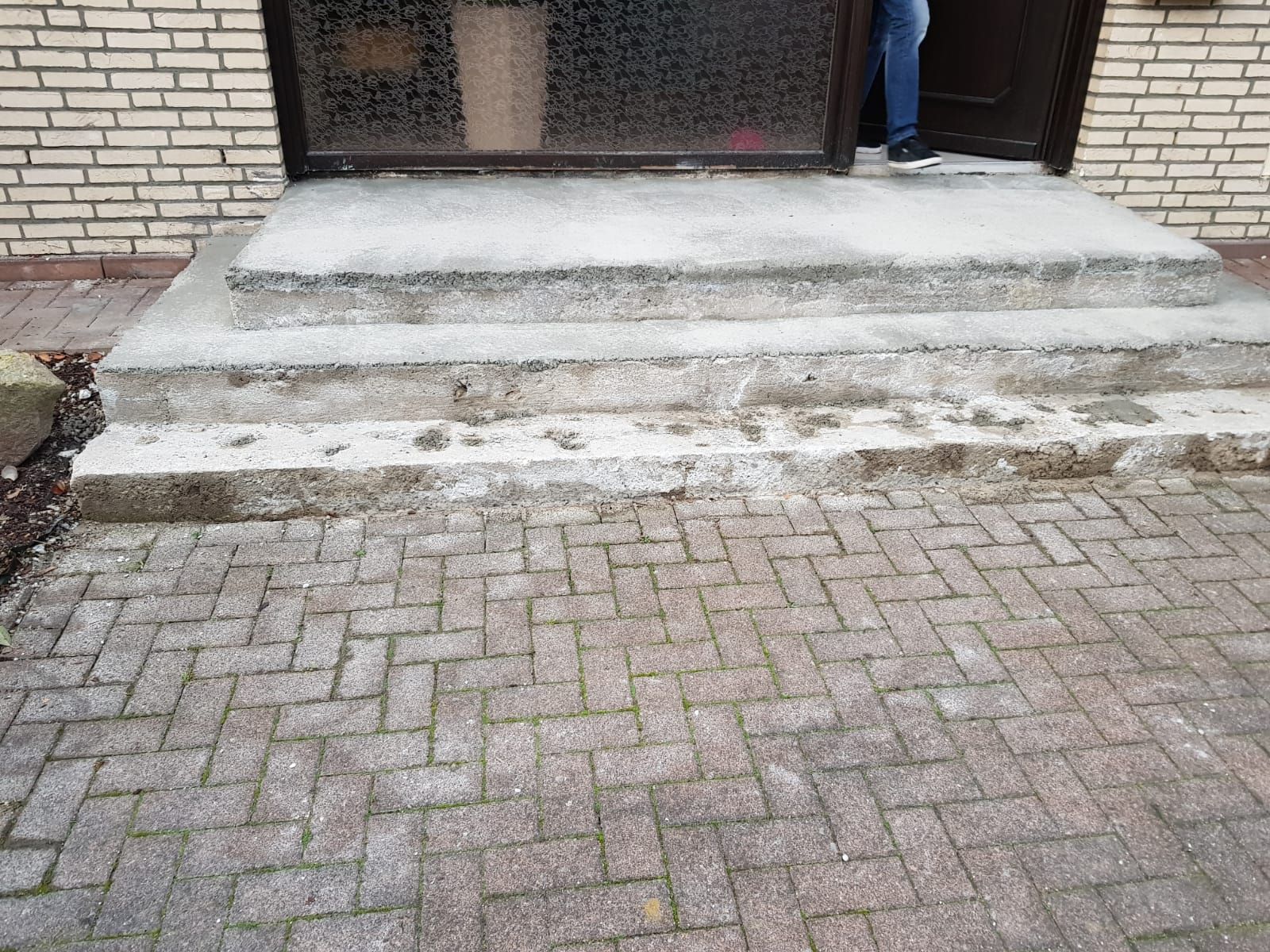 Betonstufen führen zu einer Tür auf einer gemauerten Terrasse. Im Türrahmen ist eine Person zu sehen.