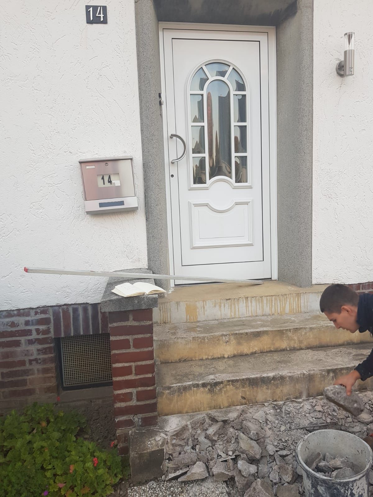 Mann repariert die Stufen eines Hauses mit weißer Tür, Briefkasten und roten Ziegelakzenten.