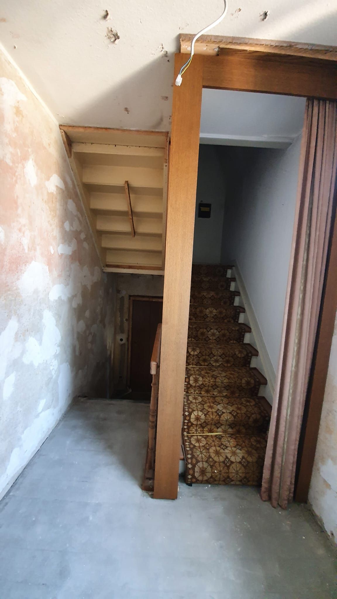 Treppenhaus mit Teppichstufen, ungestrichenen Wänden und einem Holzbalken, das zu einem dunklen Flur führt.