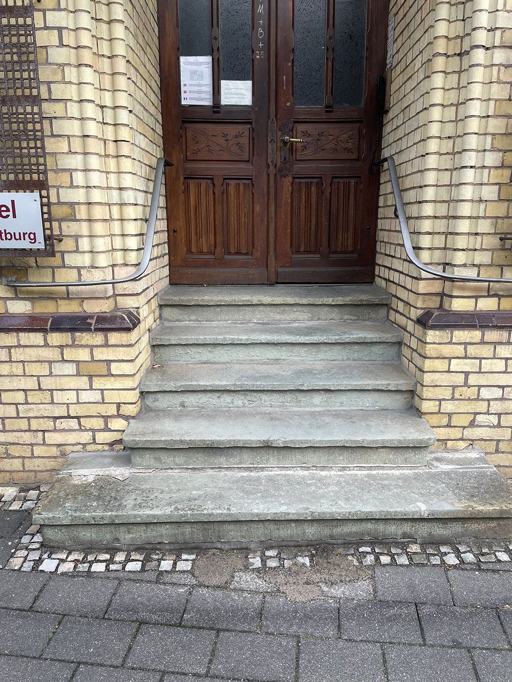 Braune Holztür mit nach oben führenden Stufen, flankiert von Ziegelsäulen und Handläufen.