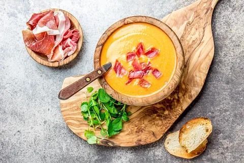 Tazón de madera con sopa de naranja, adornada con prosciutto crujiente, servida con pan y verduras.