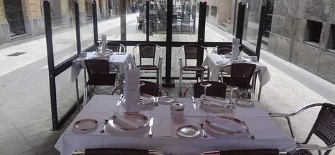 Terraza de restaurante con manteles blancos y cubiertos puestos, cerrada con cristal.
