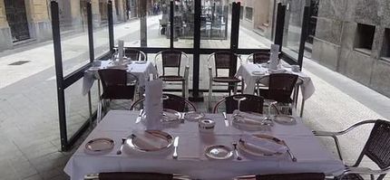 Terraza de restaurante con manteles blancos y cubiertos puestos, cerrada con cristal.