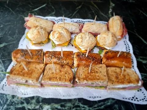 Bandeja de sándwiches variados: mini hamburguesas, sándwiches pequeños y panecillos sobre una blonda de papel decorativa.