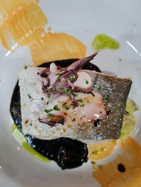 Filete de pescado sellado con pulpo, sobre salsa negra, con salsas coloridas en un plato blanco.