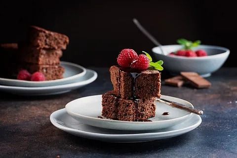 Pila de brownies de chocolate con frambuesas, salsa y menta en un plato blanco; más brownies y bayas al fondo.