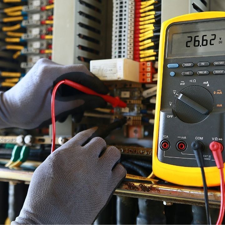 Electricista usando un multímetro para medir el voltaje en un panel de control, usando guantes; la pantalla muestra 26.62.
