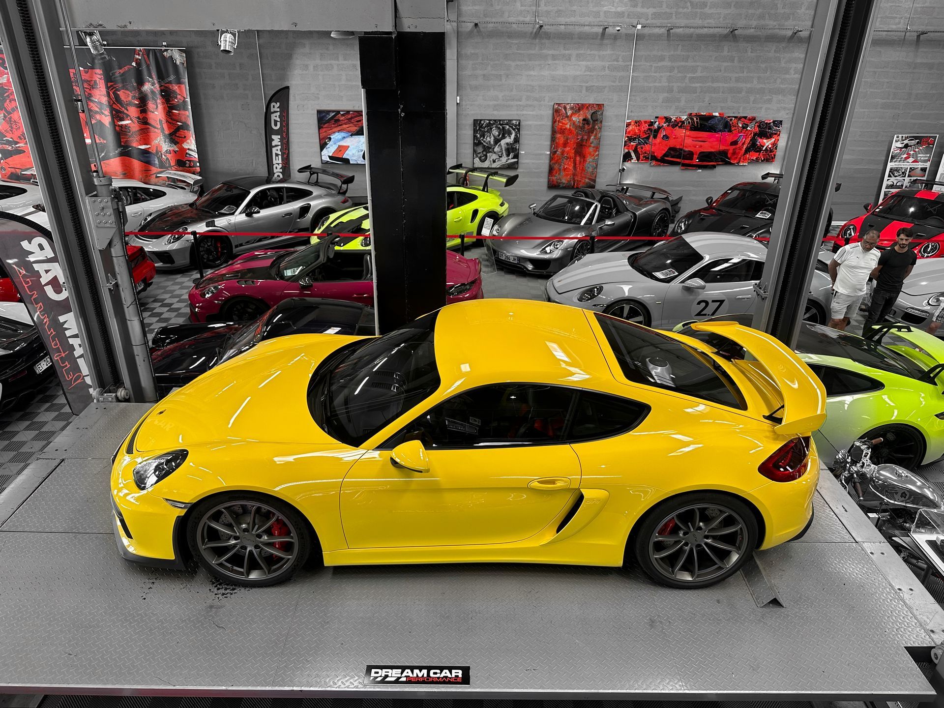 Voiture de sport jaune sur le podium d'un showroom, entourée d'autres voitures colorées et de piliers industriels.