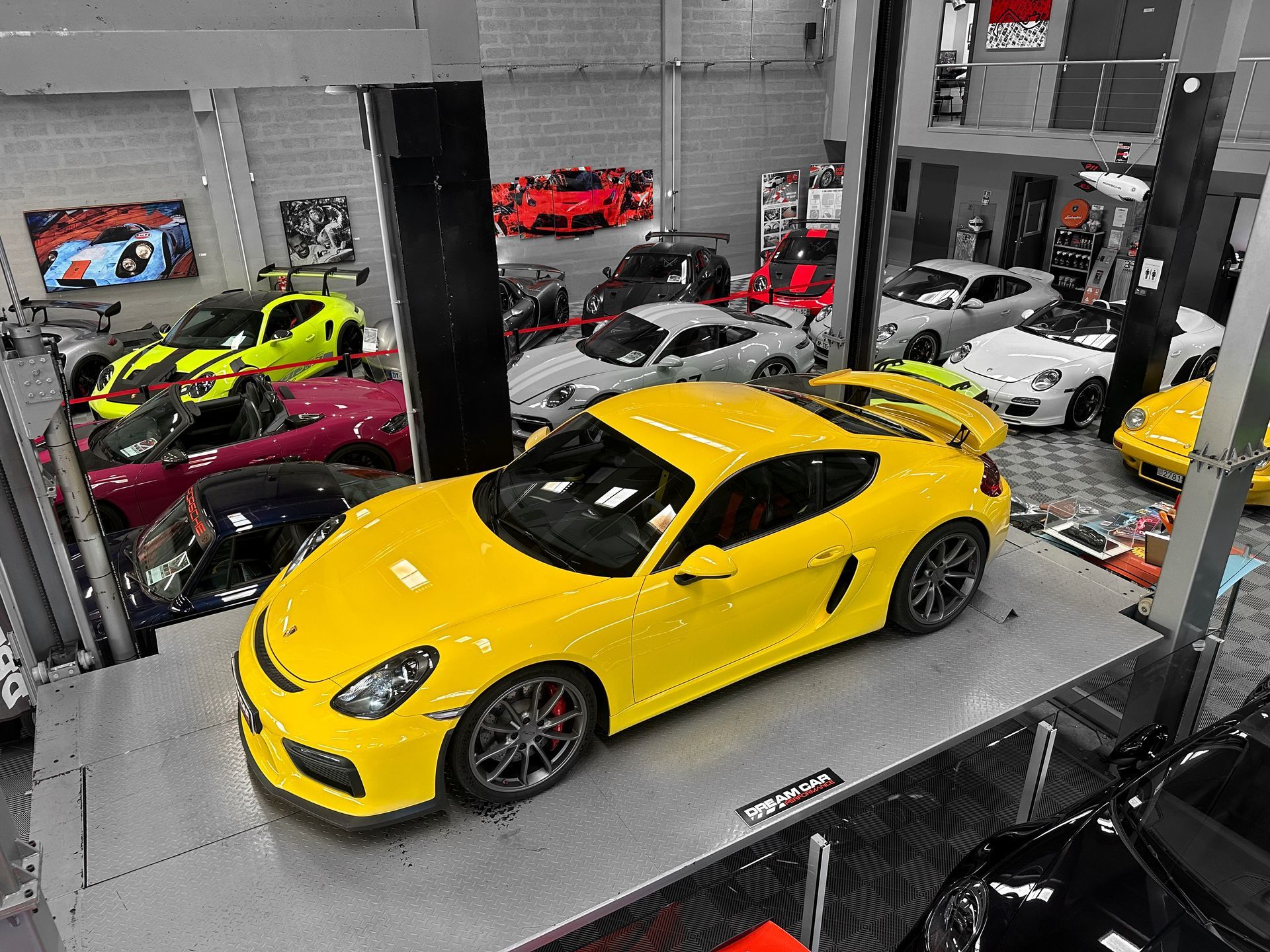Voiture de sport jaune sur le podium d'un showroom, entourée d'autres voitures colorées et de piliers industriels.