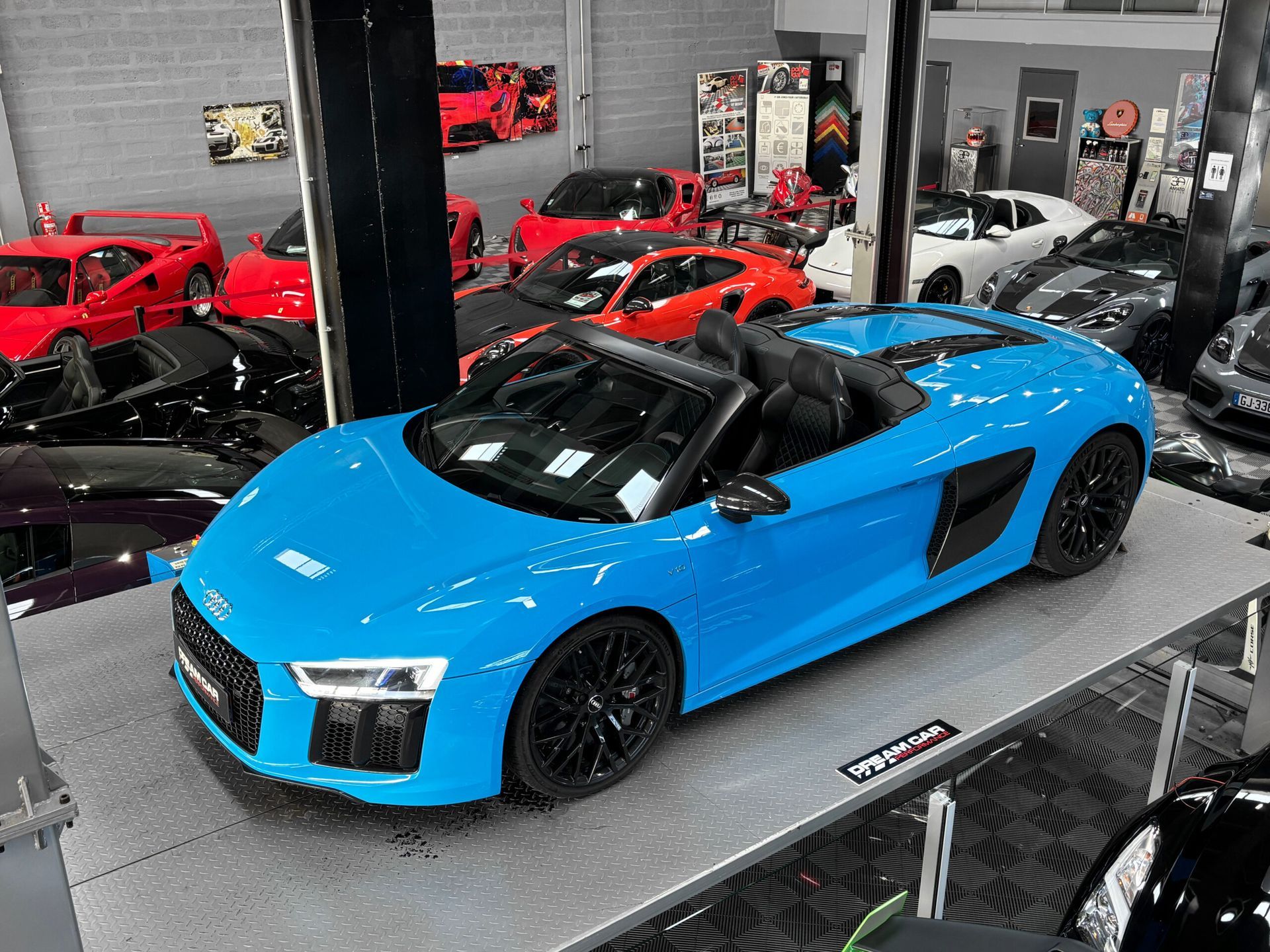 Une Audi R8 décapotable bleu vif garée sur une plateforme à l'intérieur d'un showroom rempli de voitures de luxe diverses.