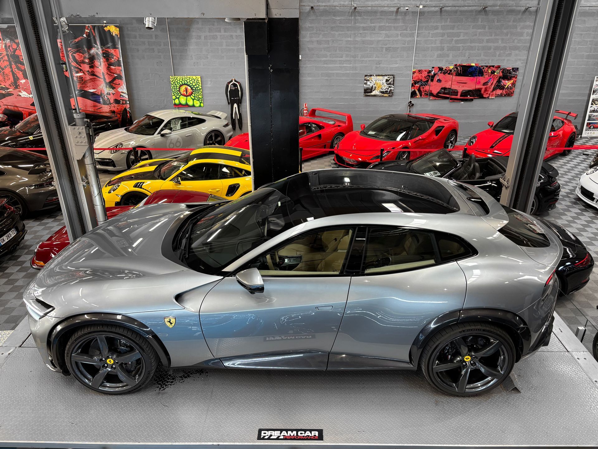Voiture de sport grise dans un showroom, avec des voitures rouges, jaunes et noires exposées derrière.