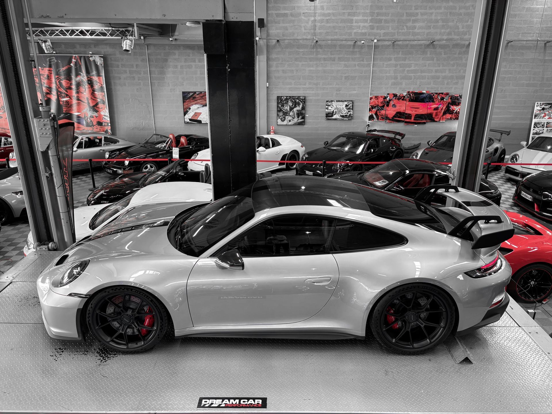 Voiture de sport argentée dans un showroom, entourée d'autres voitures et de présentoirs.
