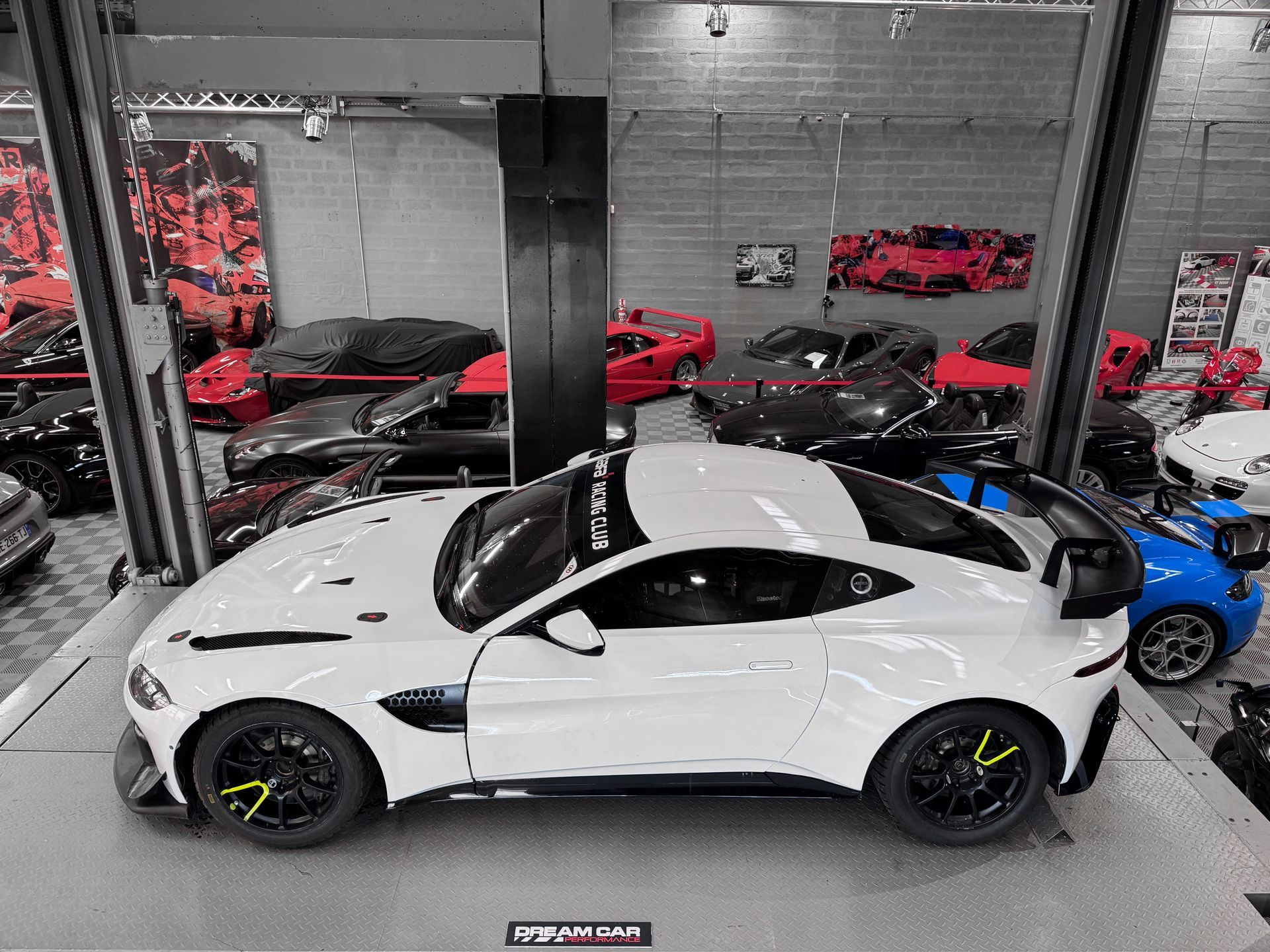 Une voiture de sport blanche est exposée sur un ascenseur de showroom à côté de voitures rouges et noires dans un showroom ressemblant à un garage.