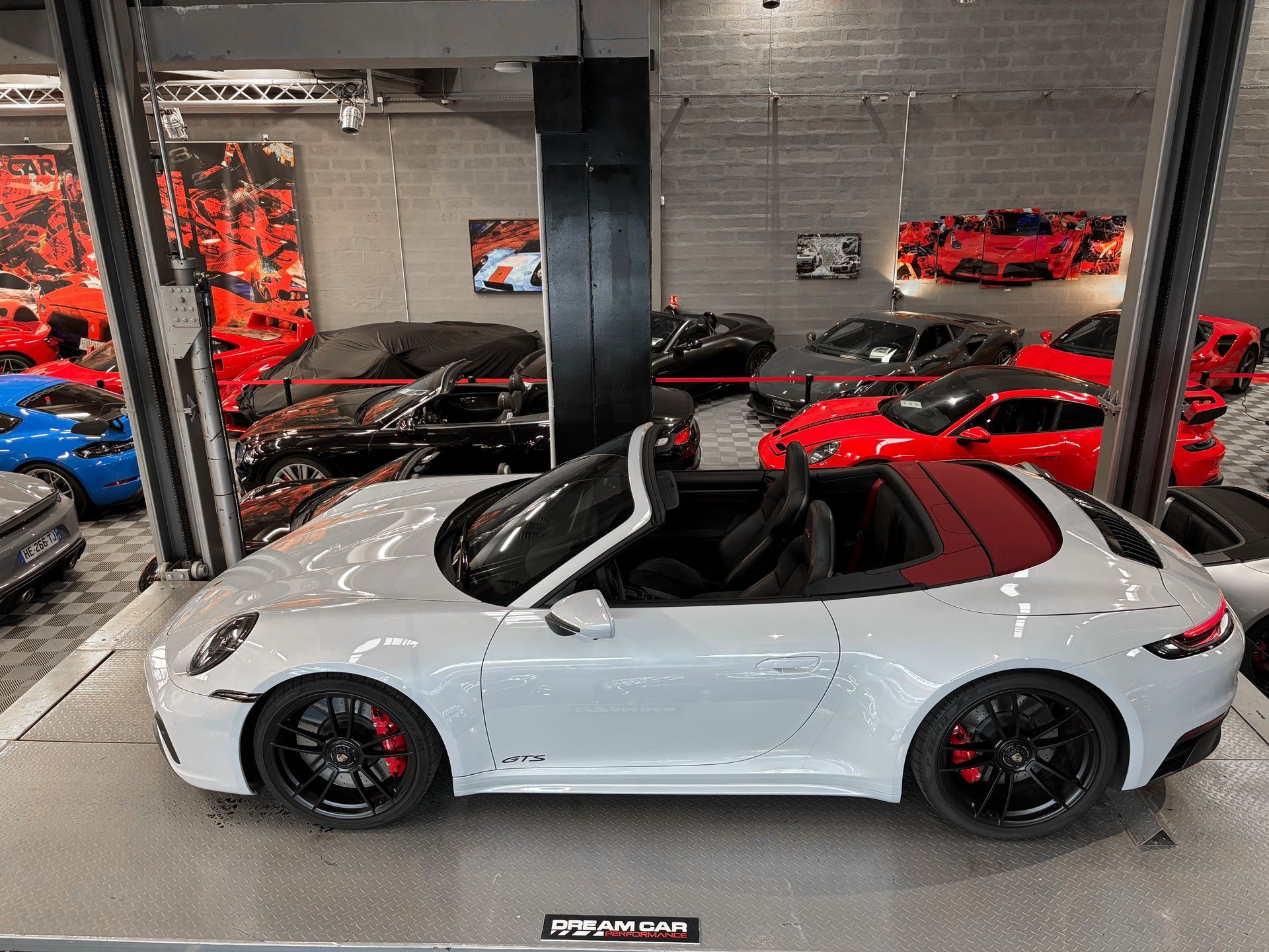Une Porsche 911 Cabriolet argentée est exposée dans un showroom de voitures de luxe, entourée de voitures de sport rouges.