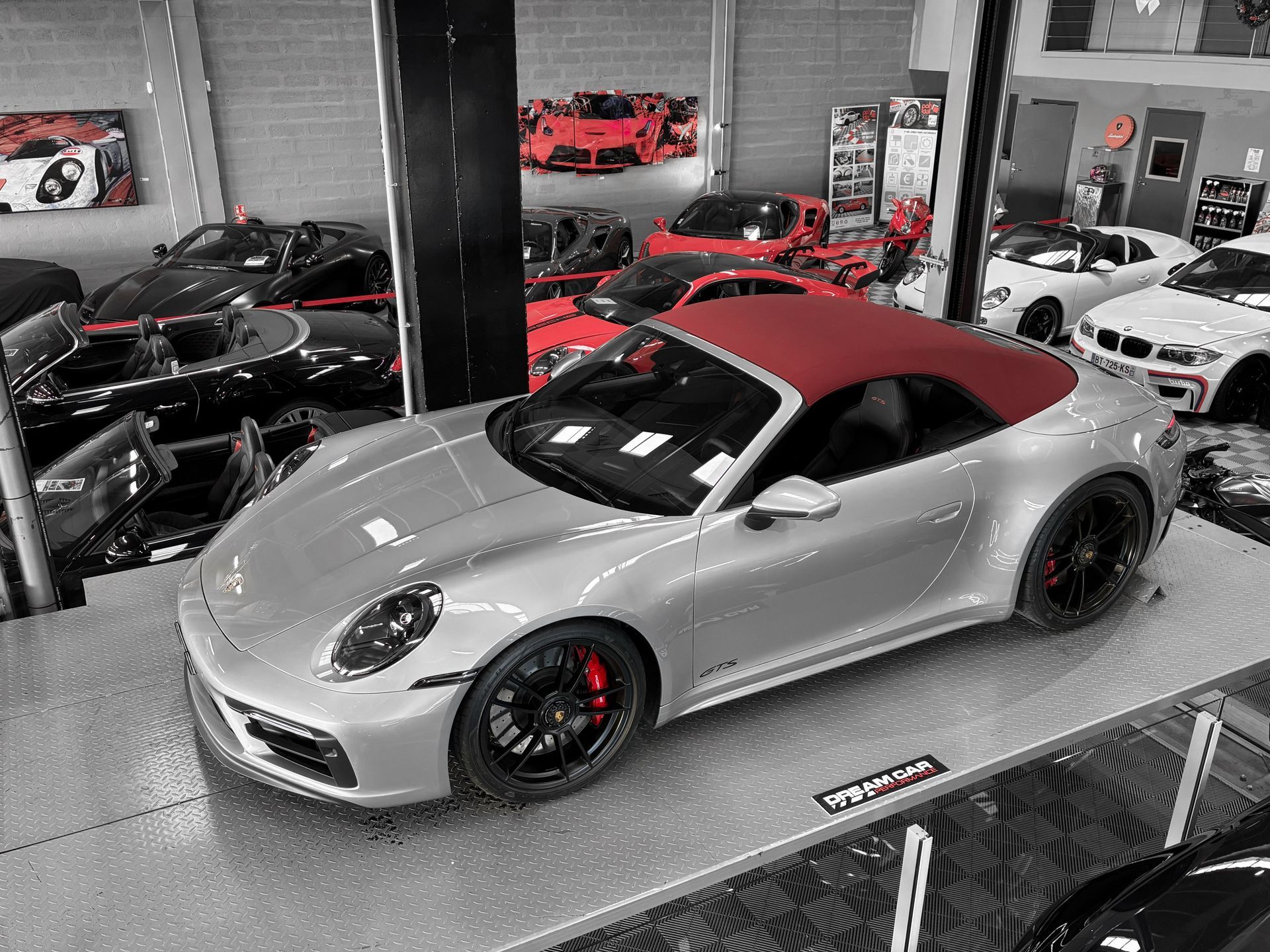 Porsche 911 Cabriolet argentée sur un plateau tournant dans une salle d'exposition, entourée d'autres voitures de sport dans une salle d'exposition de style garage