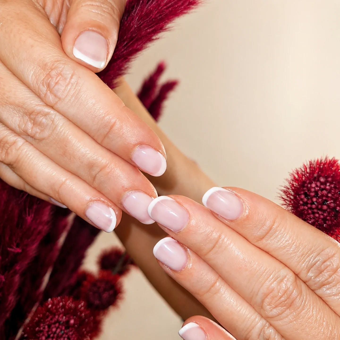 Des mains aux ongles rose pâle manucurés se touchent près de fleurs bordeaux