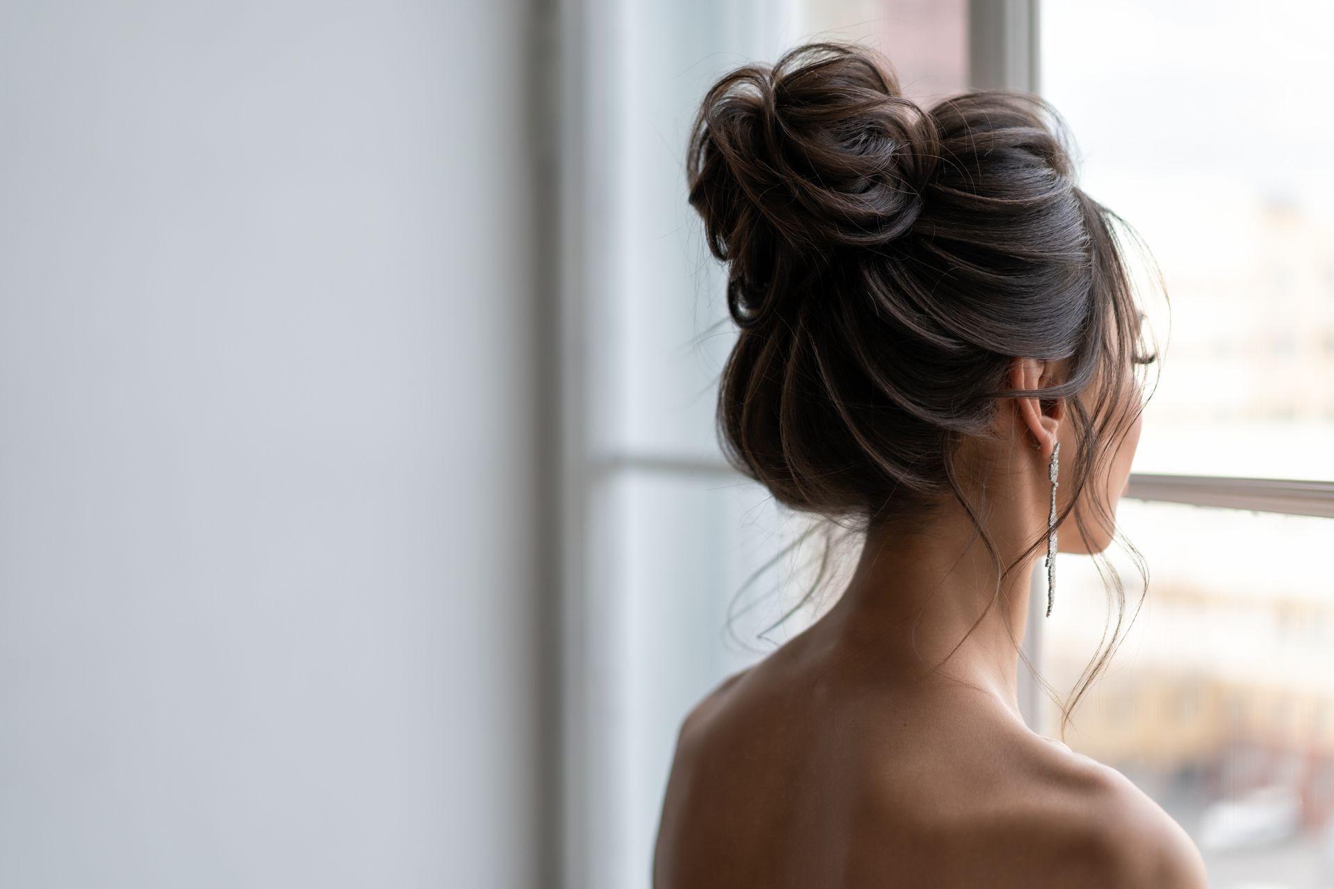 Femme aux cheveux noirs coiffés en chignon, vue de dos, debout près d'une fenêtre lumineuse