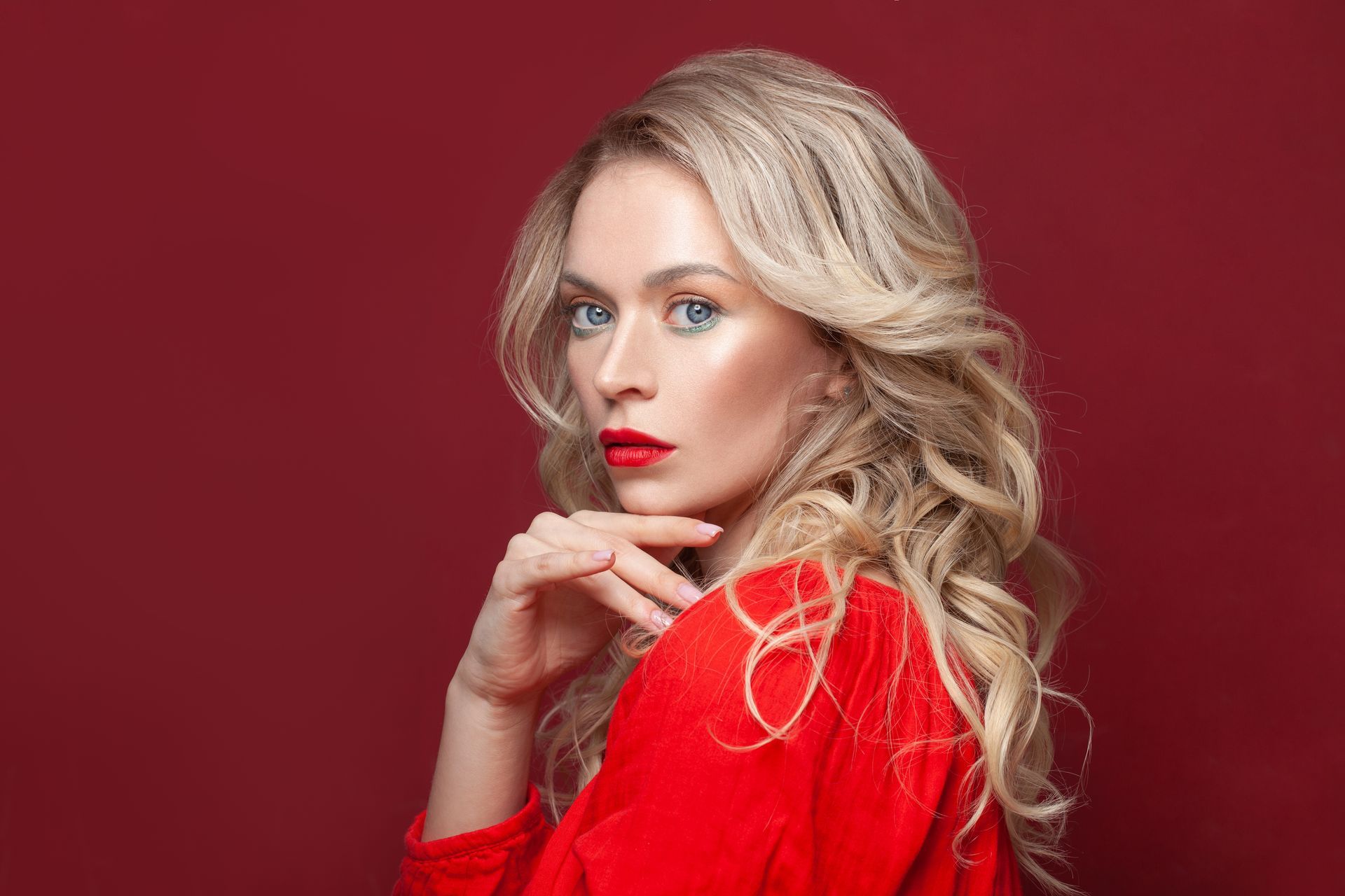 Mannequin blonde en robe rouge posant sur fond bordeaux, main sous le menton, regardant l'objectif