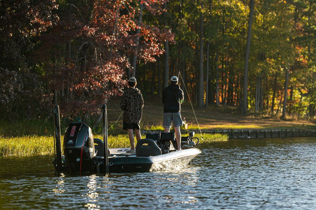 Two men fishing from a boat near a treeline. Sun is setting, casting light.
