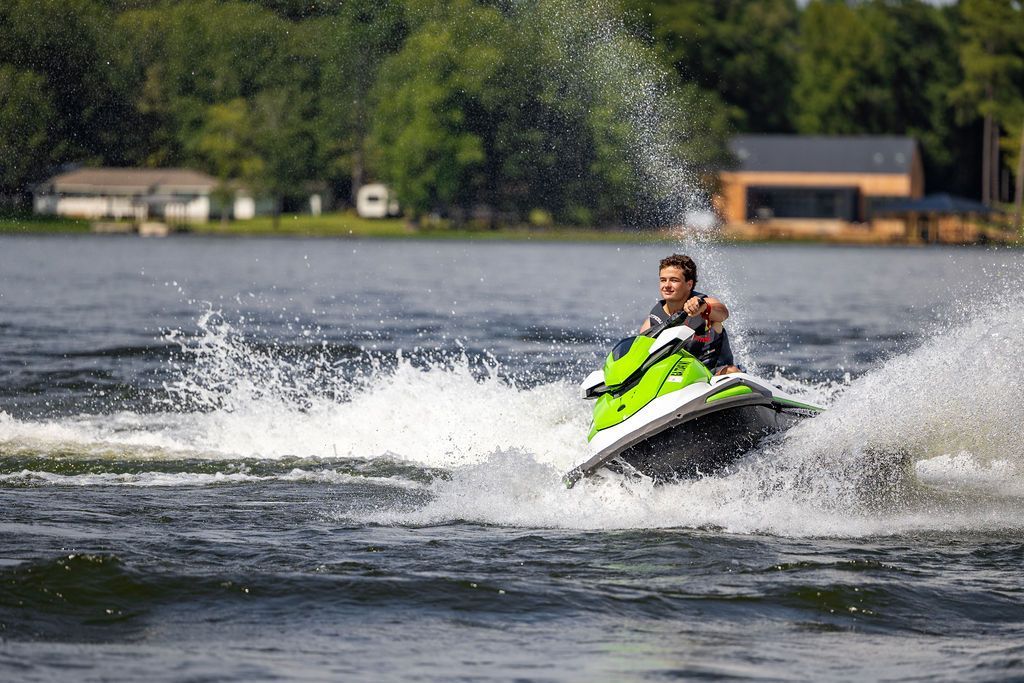 Person on a green jet ski speeding across water, creating splashes. Houses and trees in the background.