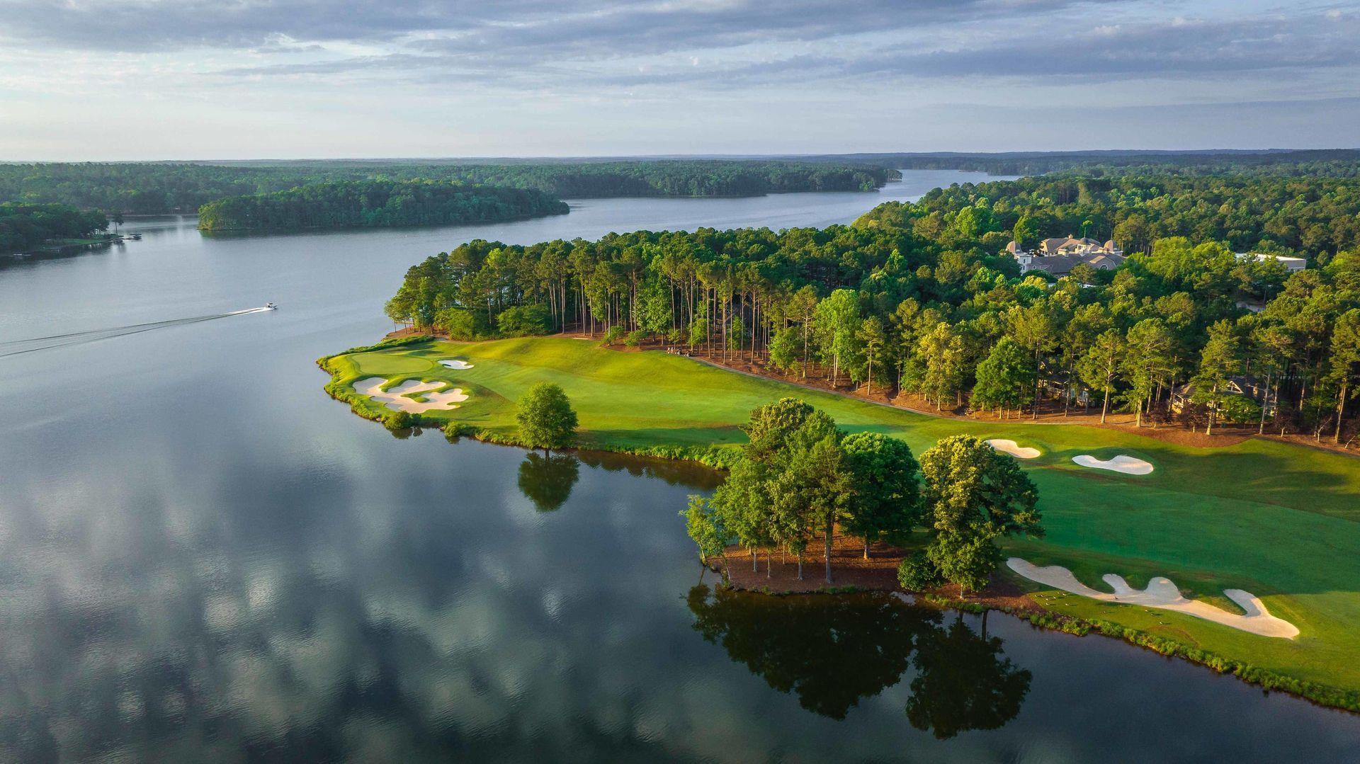 Aerial view of a golf course beside a lake. Green fairways and trees frame the water, reflecting the cloudy sky.
