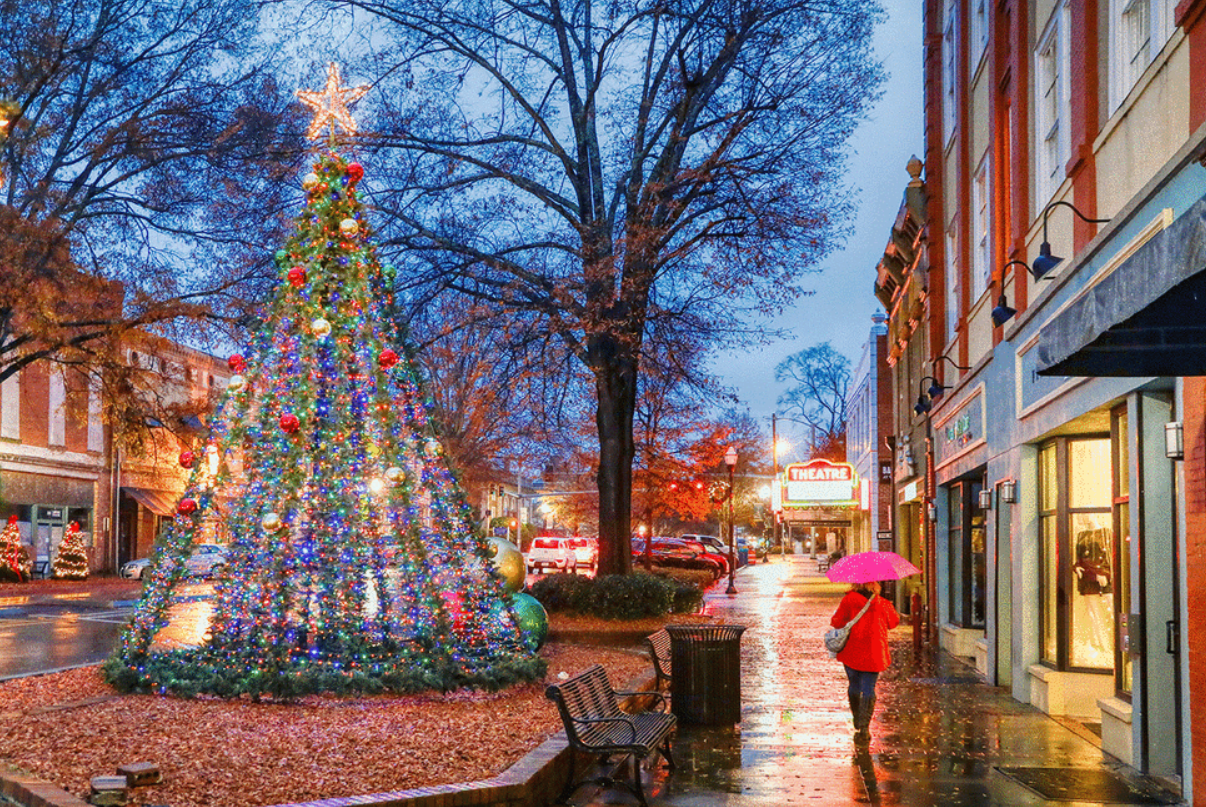 Christmas tree lit with lights, person with pink umbrella walking down wet street, downtown buildings.