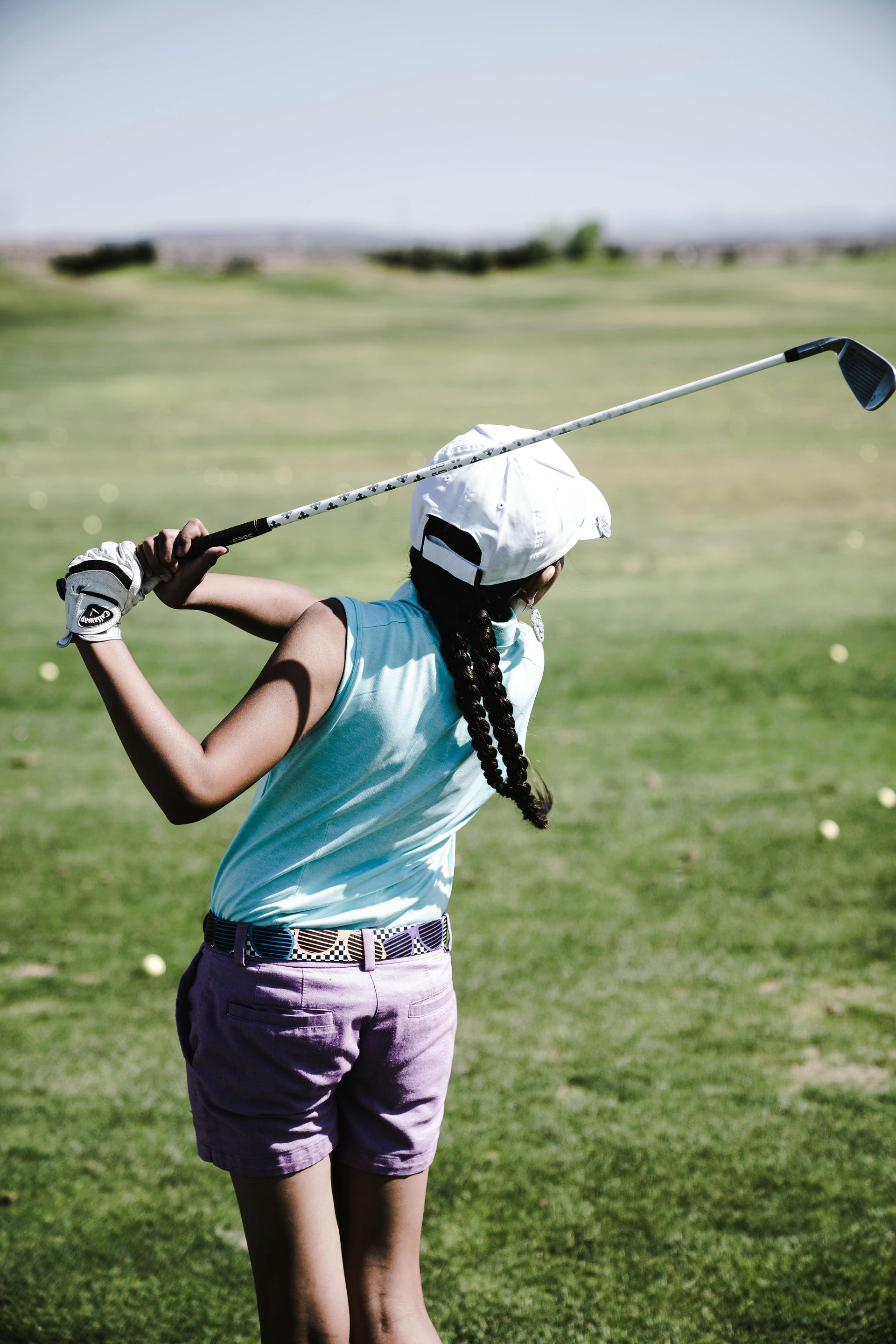 Girl in light blue shirt swings golf club on a sunny green course.