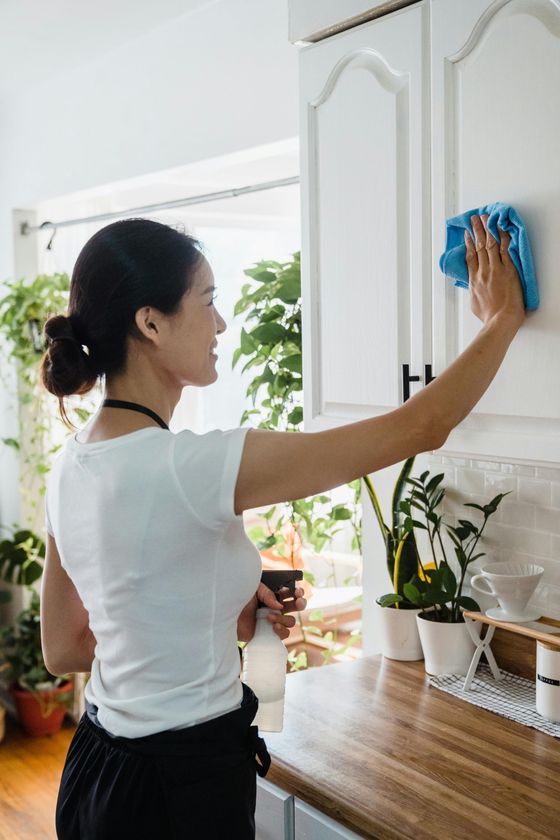 Woman cleaning a white kitchen cabinet with a blue cloth, holding a spray bottle.