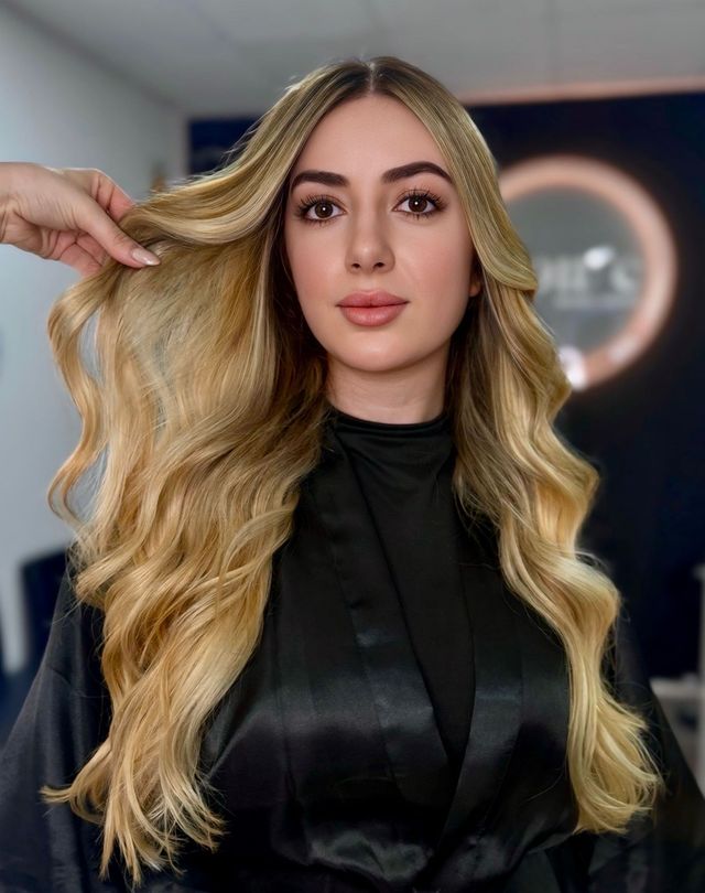Mujer con cabello largo rubio y ondulado, en un salón de peluquería, con bata negra, siendo peinada.