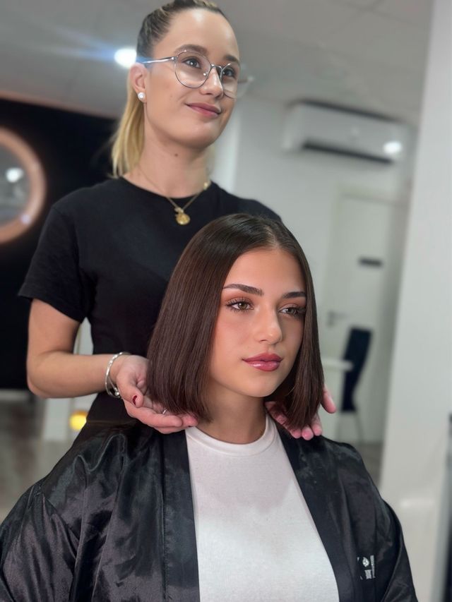 Estilista con clienta; corte bob terminado. Mujer con camisa negra y gafas, otra con top blanco, mirando a cámara.