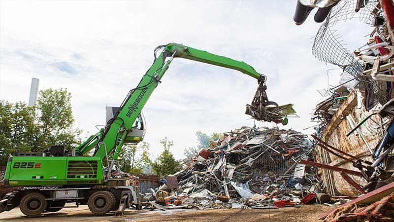 Ein grüner Bagger lädt einen Haufen Schrott auf einen Lastwagen.