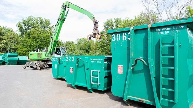 Ein grüner Kran hebt auf einem Parkplatz einen grünen Müllcontainer an.